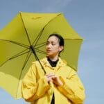 Kobieta w żółtej kurtce przeciwdeszczowej z parasolem na tle błękitnego nieba