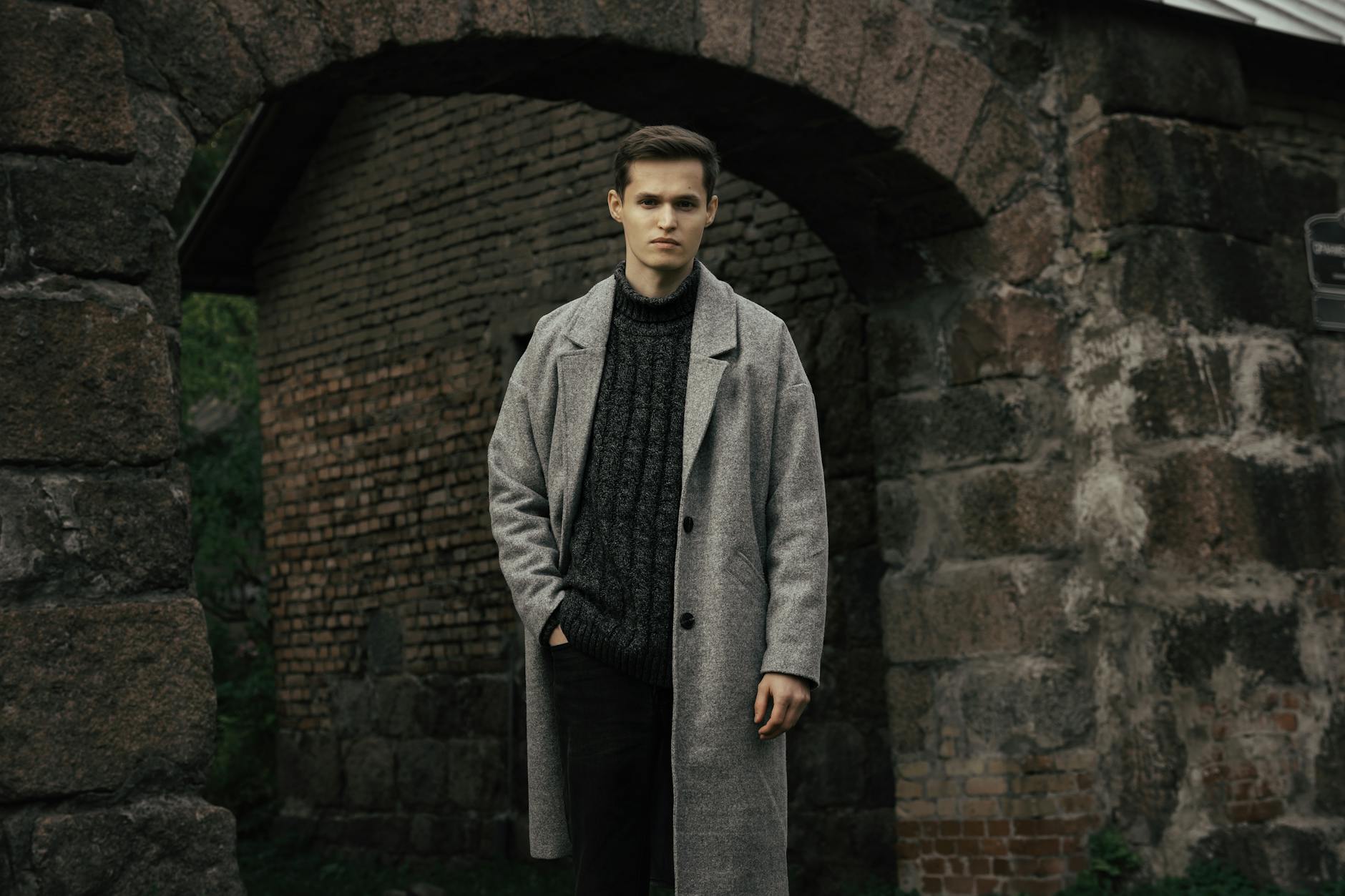 Golf, koszula czy bluza: co najlepiej wygląda pod eleganckim płaszczem na co dzień Mężczyzna w szarym płaszczu stoi przy zabytkowym łuku w mieście