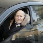 Starszy mężczyzna prowadzi auto w zimowym płaszczu, widziany przez szybę