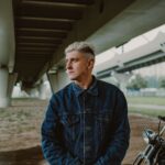 Mężczyzna w dżinsowej kurtce stoi pod mostem obok zabytkowego motocykla